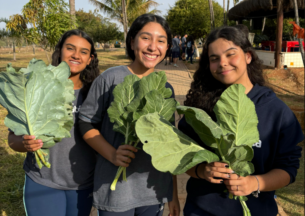 Três estudantes do Colégio segurando hortaliças que colheram na horta do colégio.