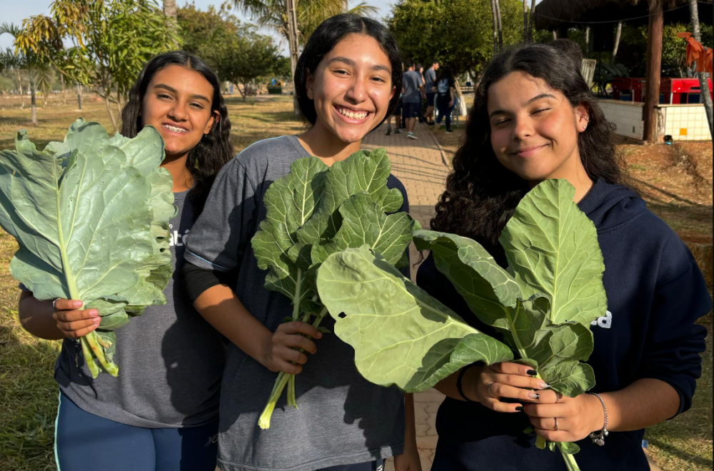 Três estudantes do Colégio segurando hortaliças que colheram na horta do colégio.